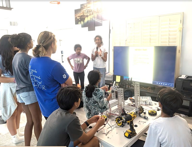 A team meeting: two members present a plan on a big screen while others assemble a drivetrain with mecanum wheels at the table